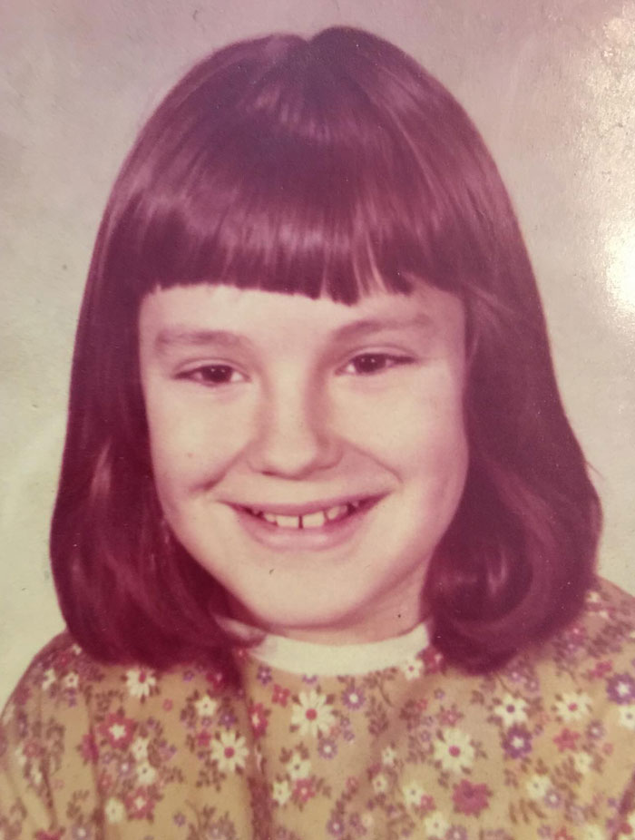 Child smiling with a childhood hair disaster featuring uneven, blunt bangs and shoulder-length hair in a floral dress.