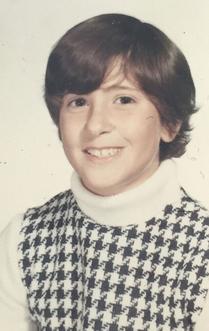 Childhood photo of a smiling young person with a hair disaster and wearing a houndstooth patterned sweater vest.