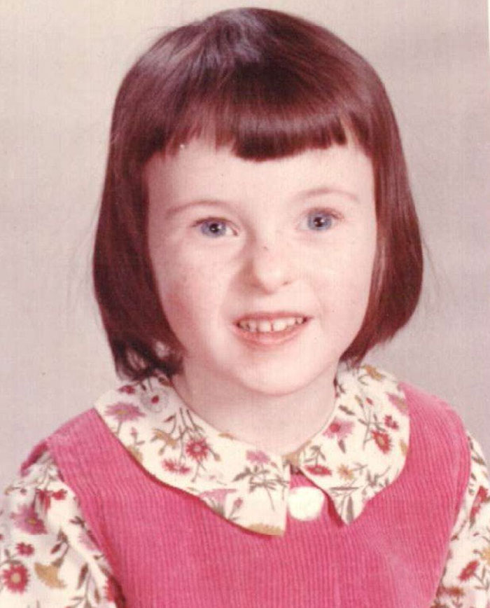 Childhood photo of a young girl with a hair disaster haircut, wearing a floral blouse and a pink pinafore dress.