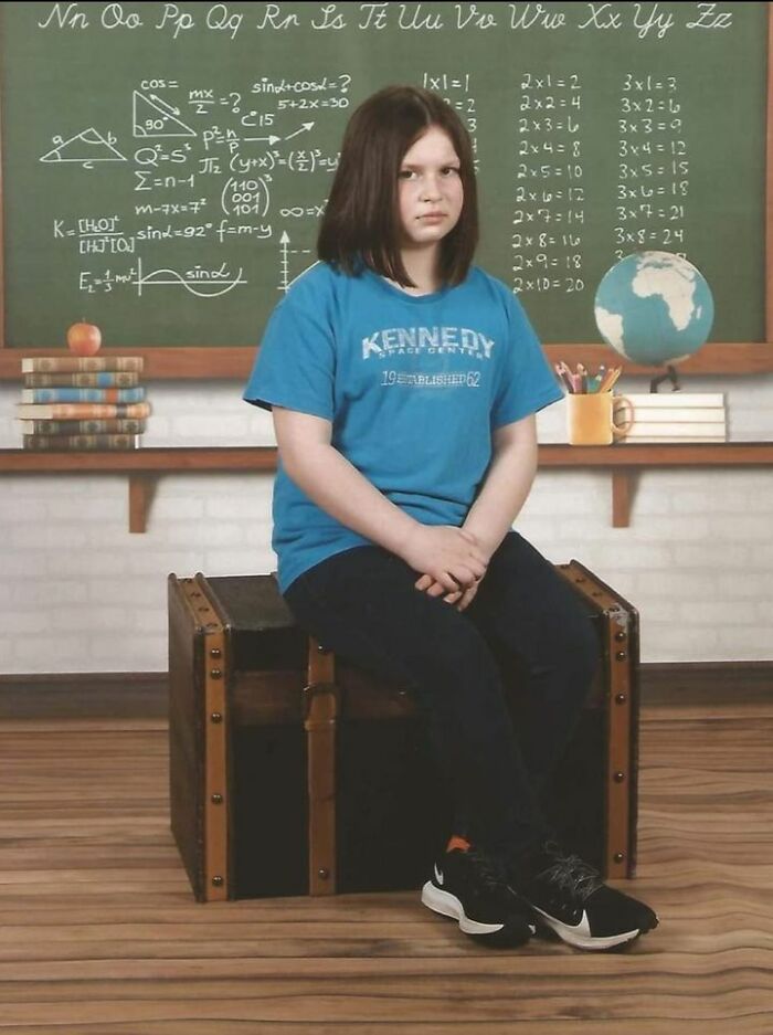 Girl wearing blue shirt with serious expression sitting on a trunk in a classroom during a school picture day fail.