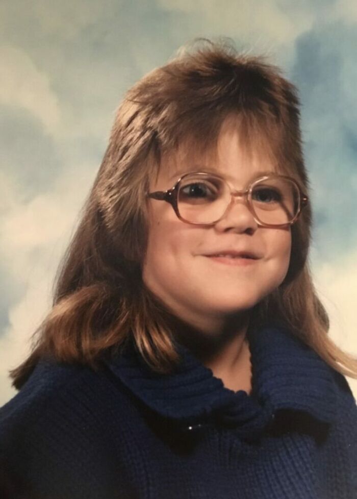 Child with glasses and mullet hairstyle posing for a funny school picture day fail with a blue sweater and a cloudy background.