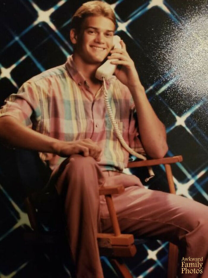 Teen boy in pastel plaid shirt and pink pants posing with a corded phone in front of a retro school picture day backdrop.