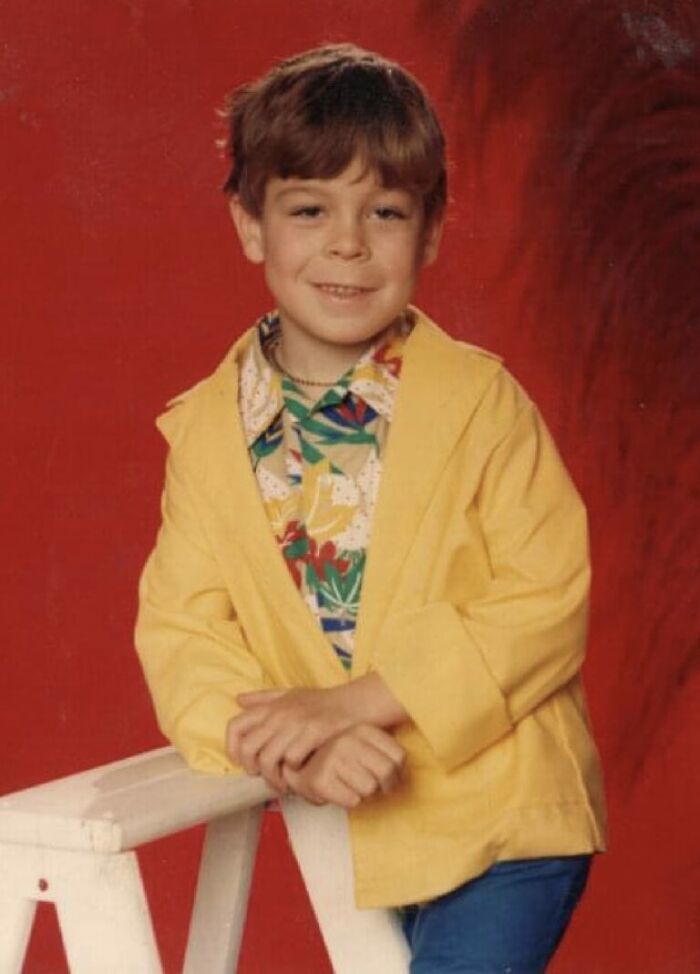 Young boy in colorful shirt and yellow jacket posing awkwardly for a funny school picture day fail against red backdrop.