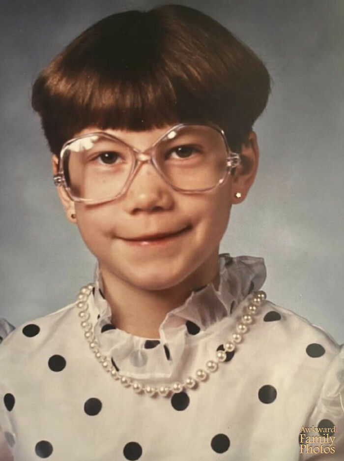 Child in oversized clear glasses, pearl necklace, and polka dot blouse posing for a funny school picture day fail.