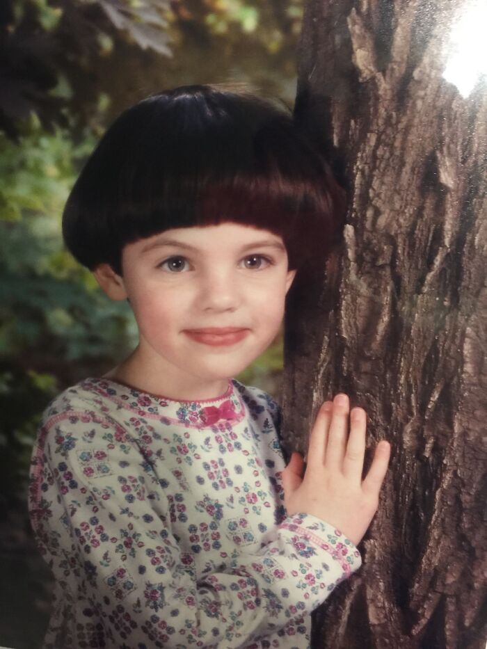 Young girl with a bowl haircut wearing a floral shirt posing by a tree in a funny school picture day fail.
