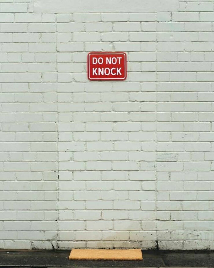Red do not knock sign on white brick wall above a doormat, a funny sign that makes people laugh or do a double take.