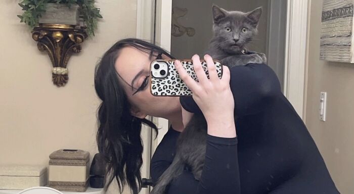 Woman taking a mirror selfie holding a gray cat with wide eyes, highlighting a funny moment of cats looking like goblins.