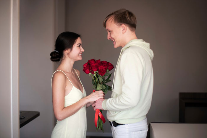 Young woman gives her boyfriend flowers, smiling at each other indoors, highlighting a moment with sexist uncle implications.