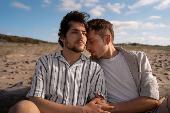 Two men sitting close on a beach, one resting his head on the other's shoulder, reflecting on household chores and work. Two men sitting close on a beach, one resting his head on the other's shoulder, reflecting on household chores and work.