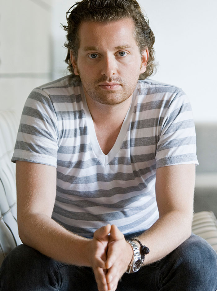 Homem gay de camisa listrada sentado com as mãos entrelaçadas, mostrando descrença e contemplação em um ambiente interno neutro.