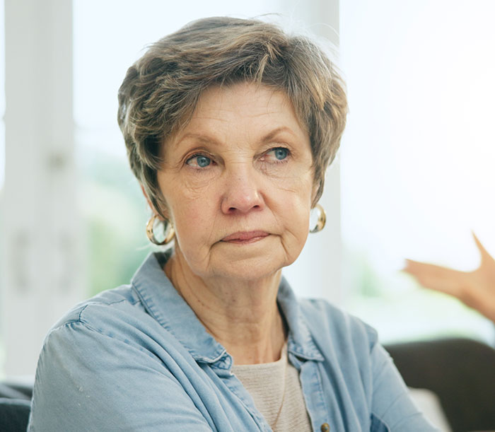 Mulher mais velha com expressão séria, vestindo camisa azul e brincos de argola dourada, sentada em um ambiente fechado perto de uma janela.