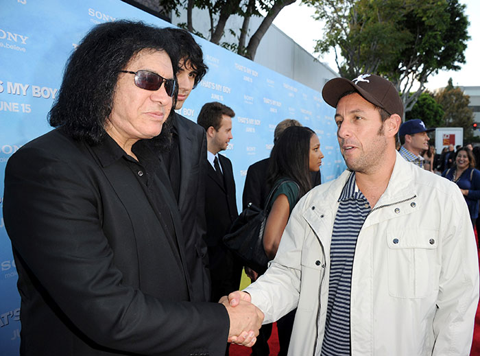 Gene Simmons and Adam Sandler shaking hands at a public event with blue promotional backdrop in the background.