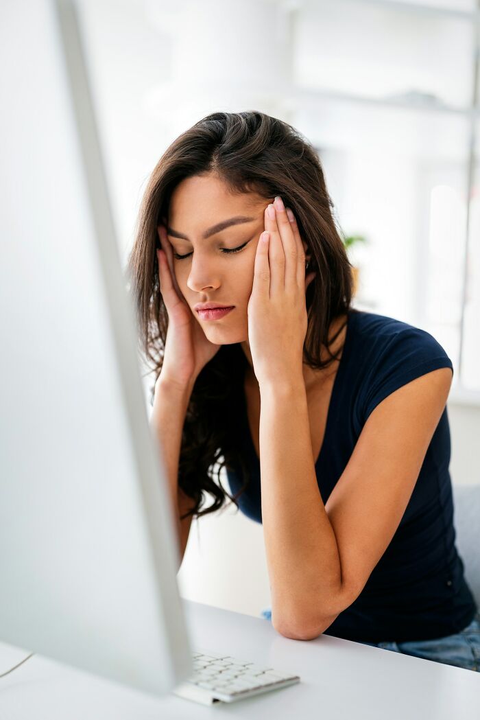 Young woman with eyes closed holding her head in front of computer, reflecting on scientific facts to blow mind today.