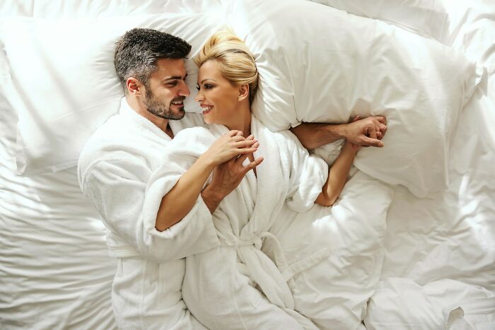 Couple in white robes lying on bed, smiling and holding hands, symbolizing forgiveness after cheating spouse issues.