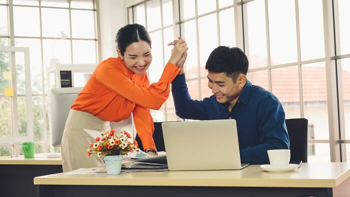 Two coworkers smiling and high-fiving at an office desk, illustrating people hit by karma after sabotage attempts.