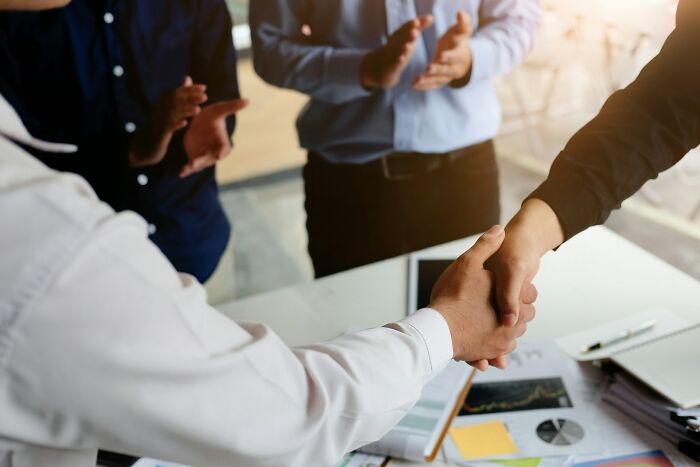 Two businesspeople shaking hands across a desk while colleagues applaud, illustrating karma in sabotage situations.