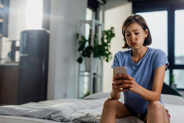 Young woman looking concerned while using smartphone in a modern bedroom, reflecting on a premium-tier zombie life hack regret.