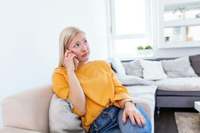 Young woman in a yellow shirt looking worried while talking on the phone in a modern living room setting.