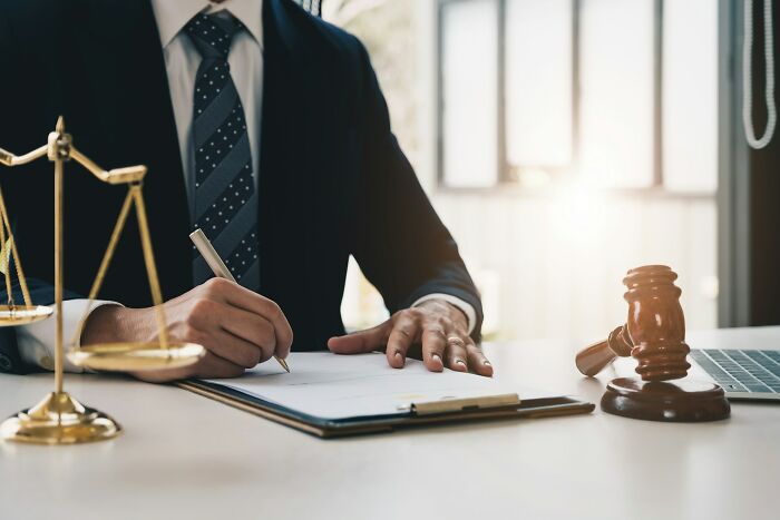 Person in a suit signing a legal document at a desk with scales of justice and a judge’s gavel nearby symbolizing life destruction.