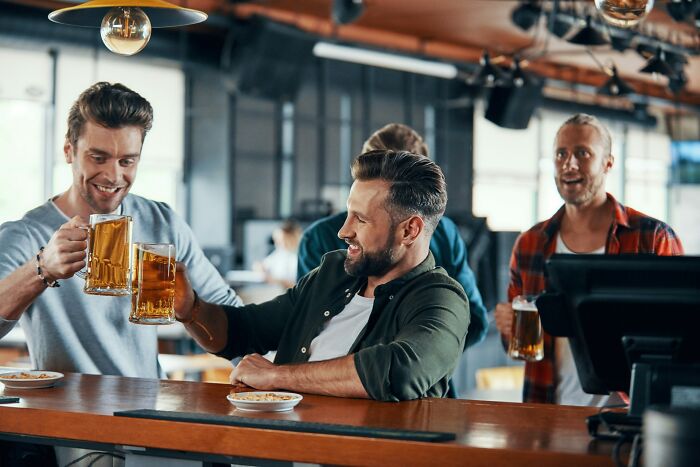 Three men clinking beer mugs at a bar, enjoying cultural things people thought were normal before visiting other countries.