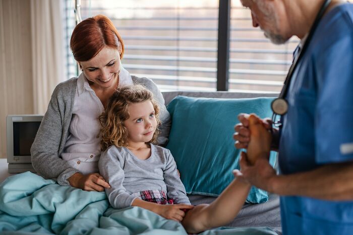 Mother and child seated on a bed while a healthcare professional in scrubs checks the child's arm carefully.