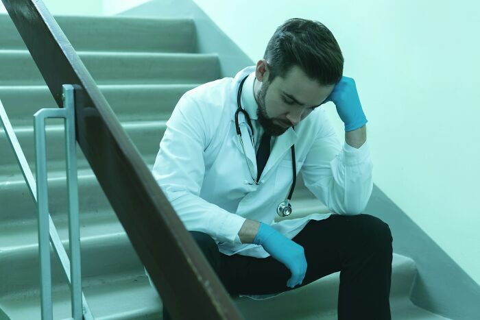 Male medical staff sitting on stairs in ER, wearing a white coat and gloves, appearing stressed and overwhelmed.