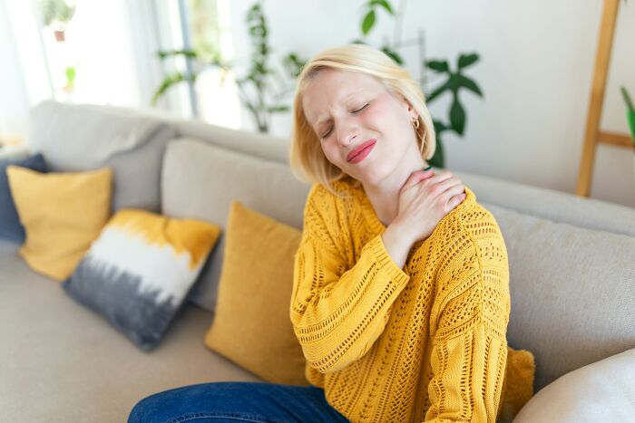 Young woman in a yellow sweater sitting on a couch, holding her neck in pain, indicating malfunctioning brain symptoms.