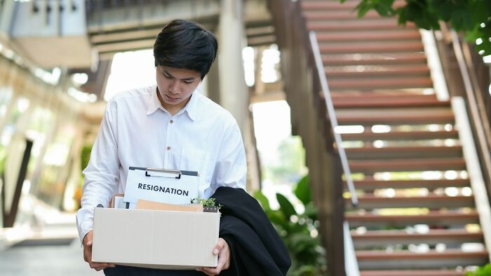 Young man holding a box with resignation papers, leaving office after karma backfired during sabotage attempt.