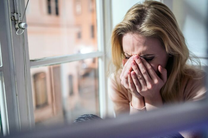 Woman sitting by a window covering her face, reflecting emotional pain while trying to forgive a cheating spouse.