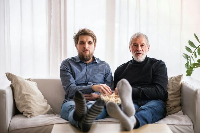 Two men sitting on a couch with popcorn, illustrating weird and unhinged family traditions at home.