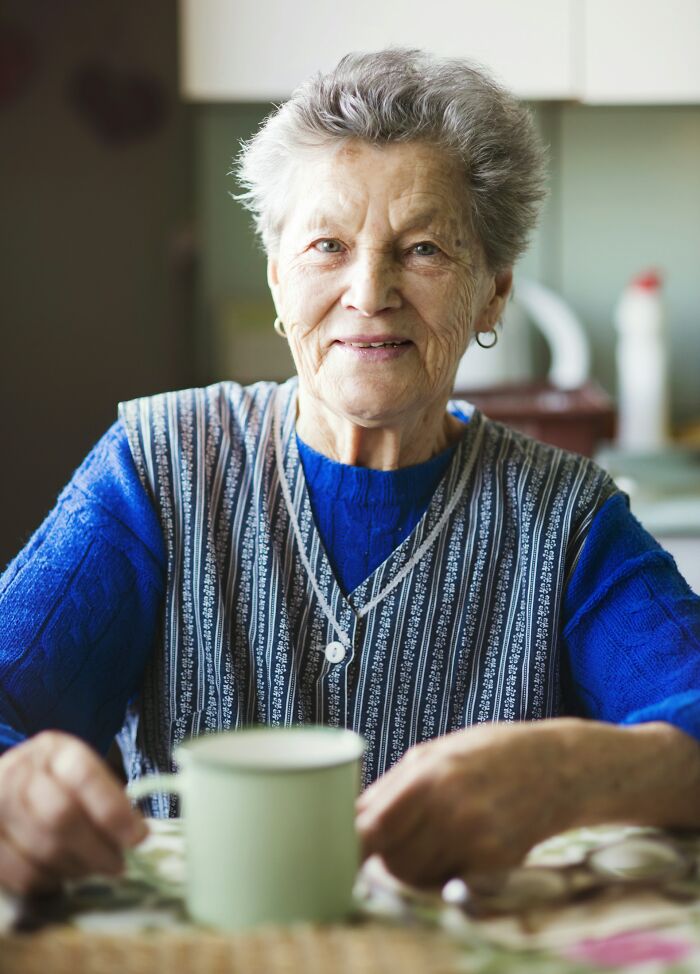 Elderly woman smiling warmly while sitting at a table, holding a cup, evoking stories of payback moments.