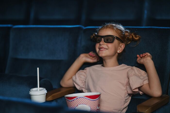 Young girl wearing 3D glasses at the movie theater with popcorn and drink, showing wholesome dad support moments.