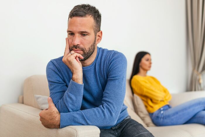 Man in blue sweater looking thoughtful on couch while woman in yellow sweater sits upset in background, forgiveness and cheating spouse concept
