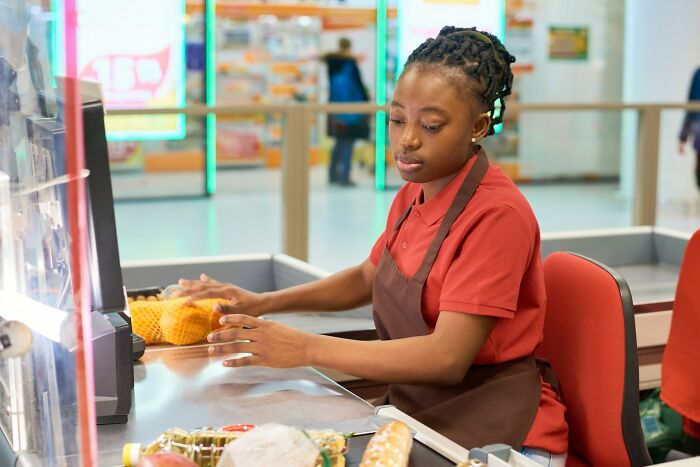 Young woman cashier in red shirt and apron working at grocery store checkout, demonstrating confident and amazing women strength.