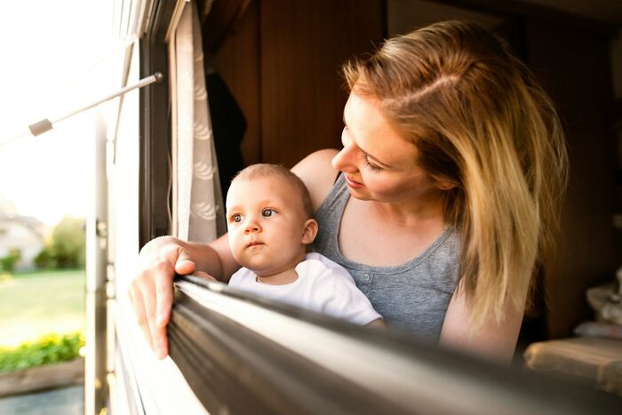 Woman with blonde hair holding a baby by an open window, looking outside on a sunny day, reflecting on age and youth.