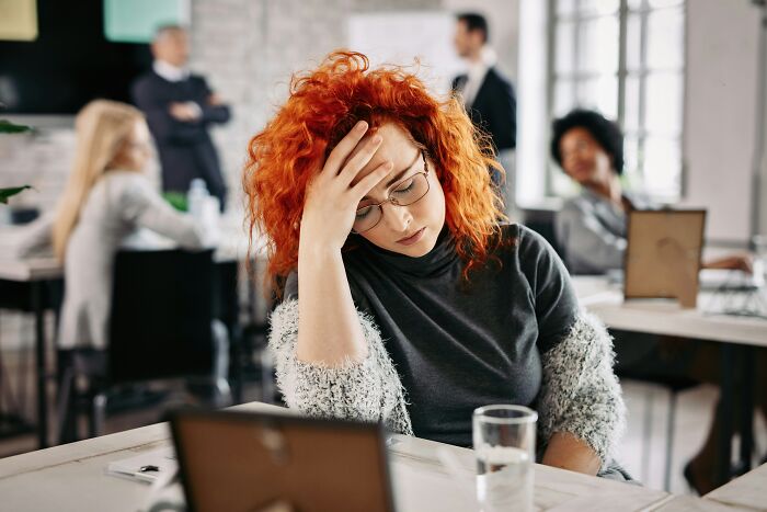Red-haired woman holding her forehead in frustration at office, showing regret after opening her mouth in a work setting.