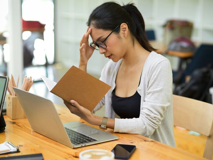 Young woman reading a book and working on a laptop, reacting with a thoughtful stare related to Gen Z stare debate.