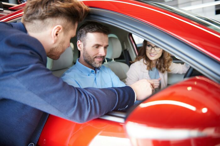 Man and woman sitting inside red car while a salesman explains features, illustrating things people heard and prayed were a joke.