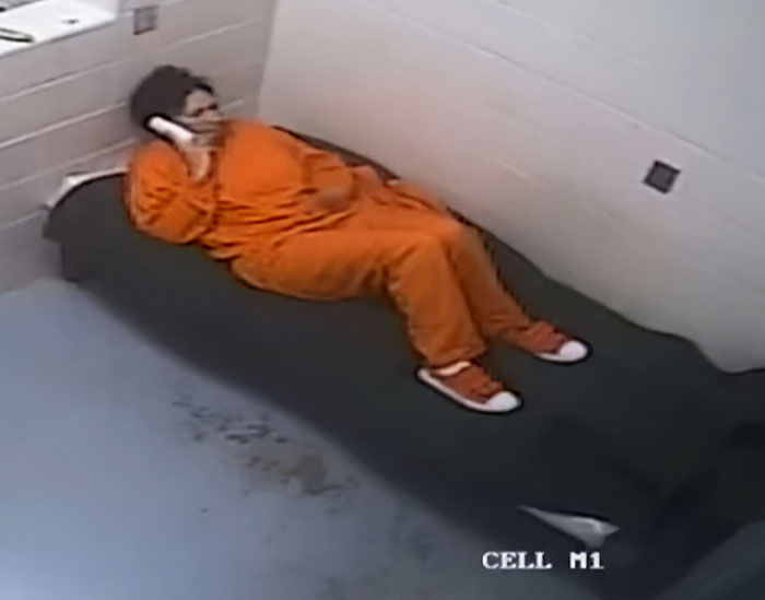 Ghislaine Maxwell in an orange prison uniform lying on a bed talking on a phone in a jail cell labeled CELL M1.