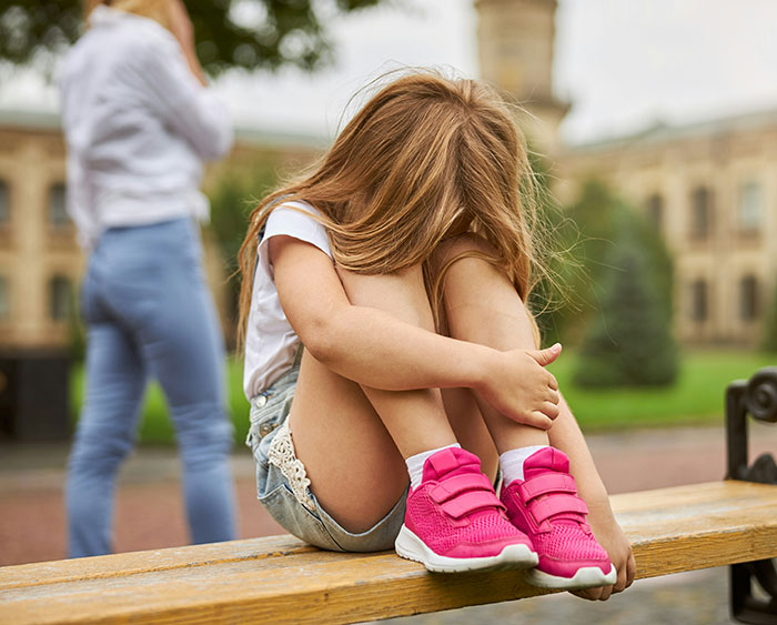 Young girl sitting alone on a bench, hugging knees, with an adult standing in the background at an outdoor public area.