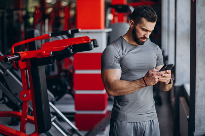 Man in gray workout clothes at gym looking at phone, relating to boyfriend lets girlfriend use apartment to catch up with ex.