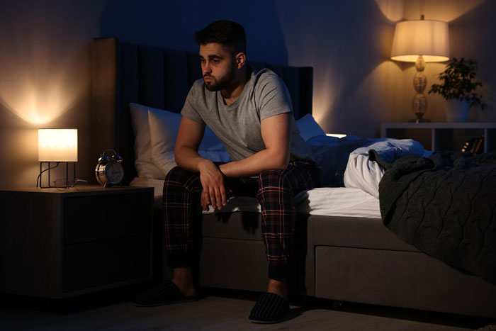 Man sitting on bed in apartment looking upset after learning about girlfriend's ex visiting as old friend at night.