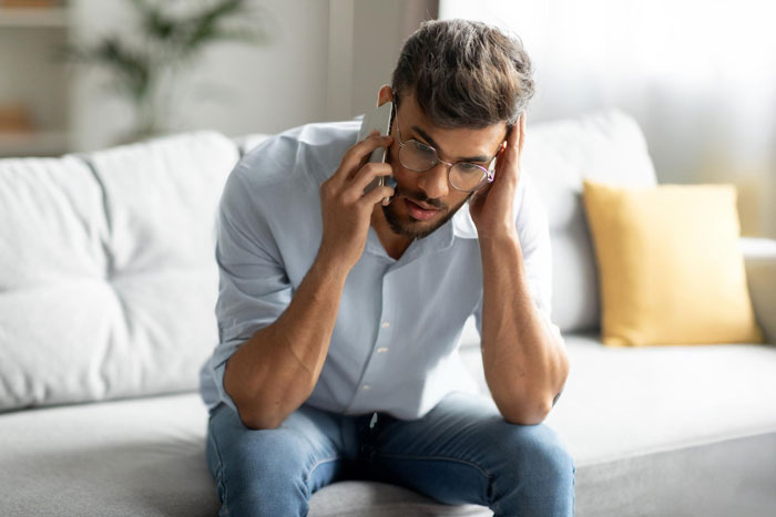 Young man in glasses sitting on a couch, looking stressed while talking on the phone in a modern apartment.