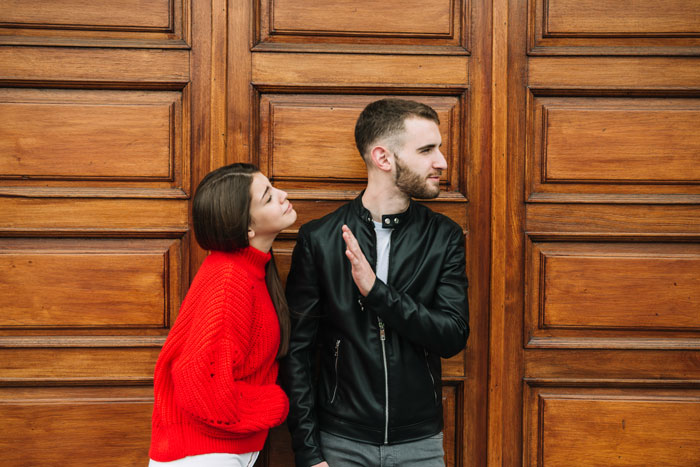 Man dumps girlfriend over open relationship request, looking away as woman tries to reconnect against wooden door background.
