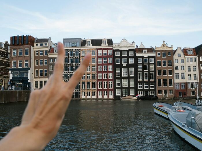 Hand showing peace sign in front of traditional canal houses and boats, reflecting vanished truths in home countries.
