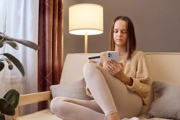 Young woman holding a credit card and smartphone at home, showing the struggle of entitled freeloaders treating friends like an ATM.