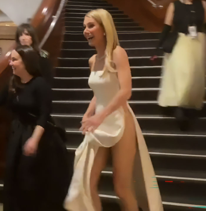 Woman in a white racy dress laughing backstage on stairs, capturing a moment related to Oscars dress mishap and aide’s reaction.