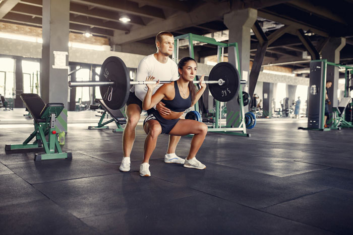 Couple training in gym, man spotting woman lifting barbell, focused on fitness and strength exercises.