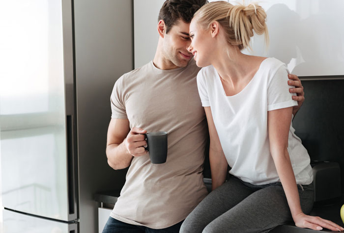 Man without shirt holding a mug, sitting close and smiling with a woman in casual clothes in a kitchen setting.