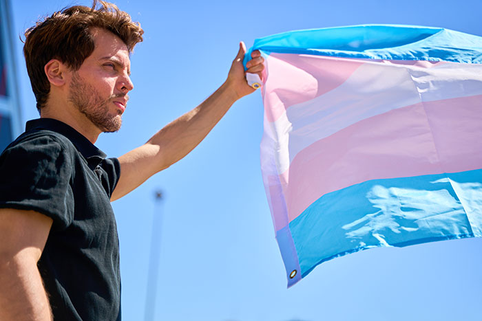 Man holding a transgender pride flag outdoors, symbolizing a man who now lives as a man in the US.